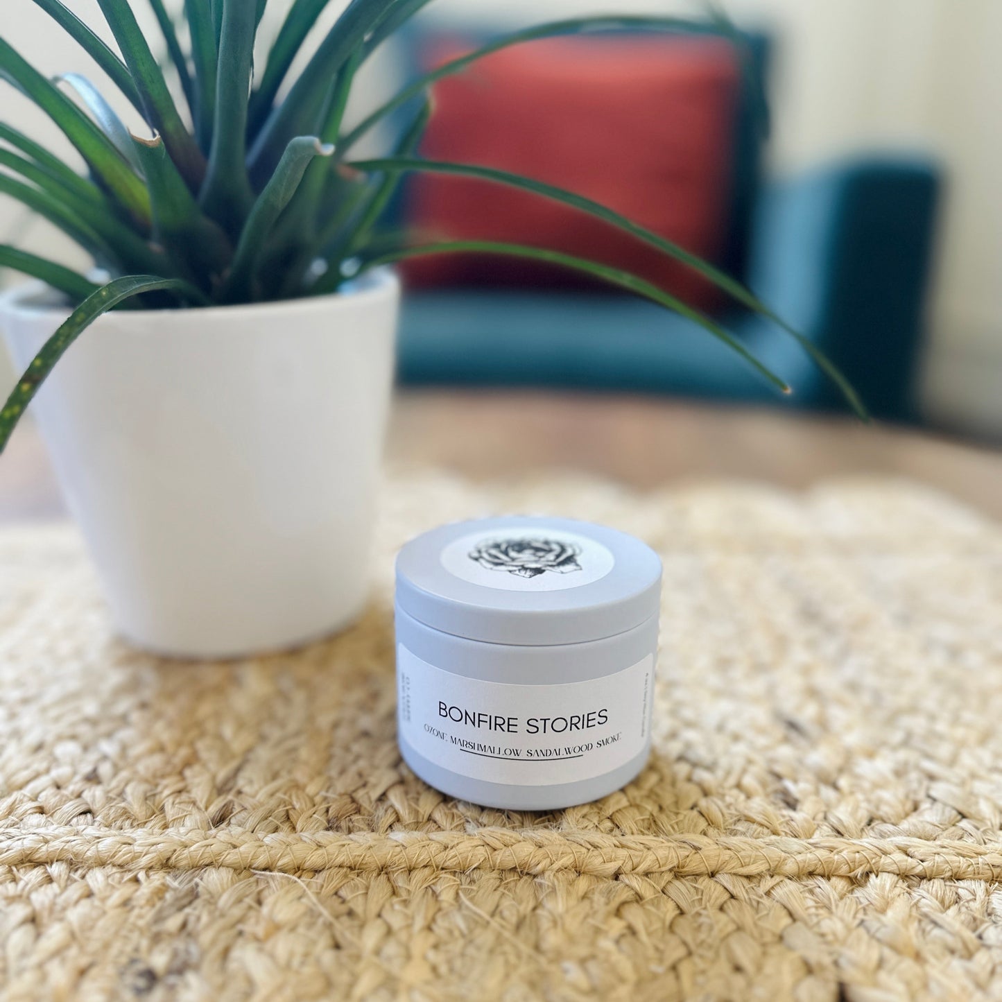Small white jar with 'Bonfire Stories' branding on a textured surface with a plant in the background